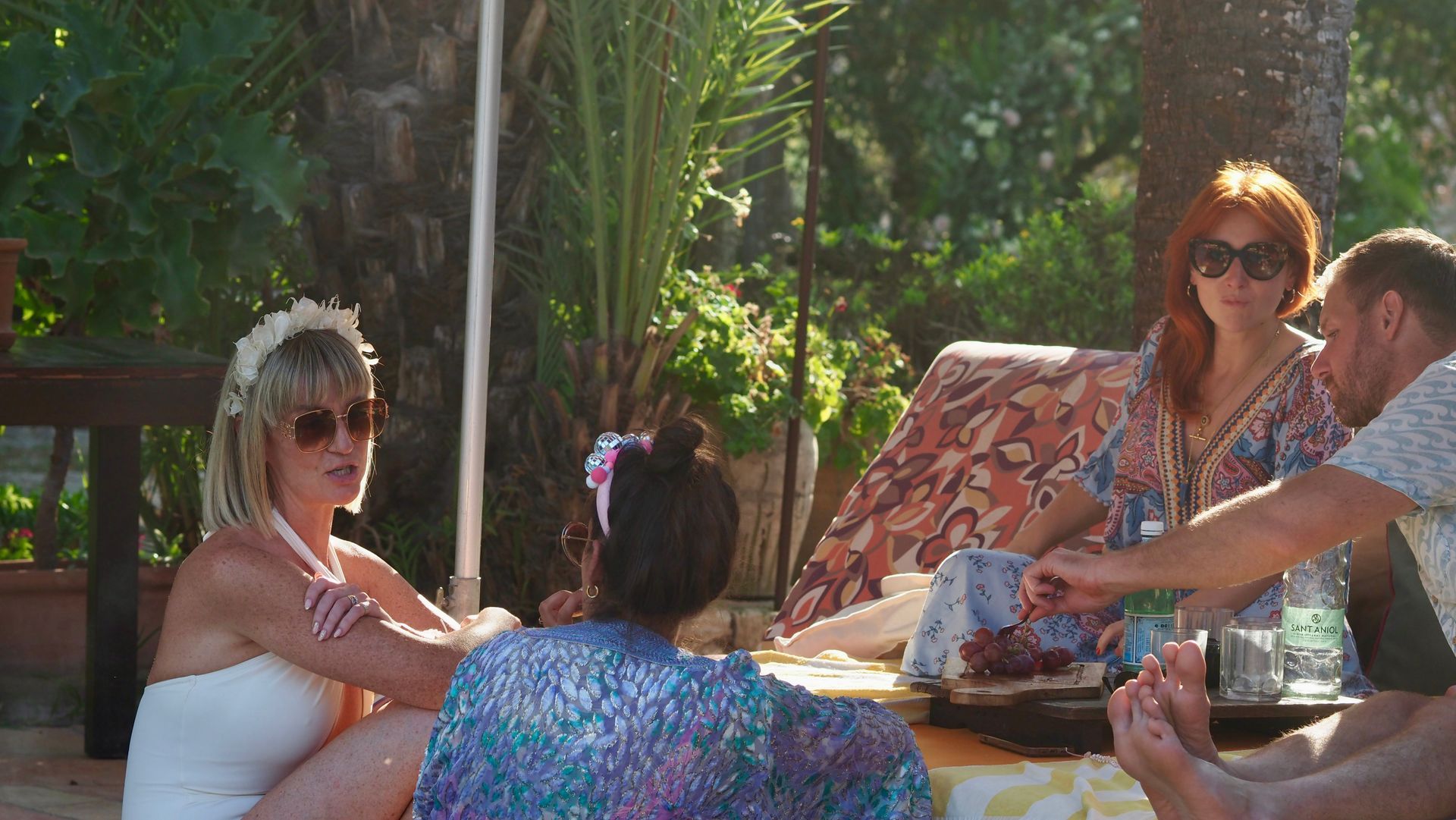 People seated around a table outdoors in a sunny garden, chatting and relaxing under palm trees.