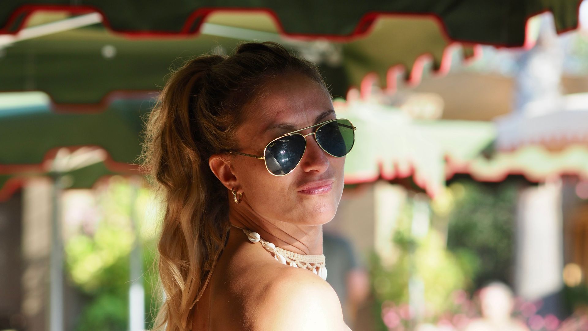 Woman wearing sunglasses turns back toward the camera under a striped outdoor canopy.