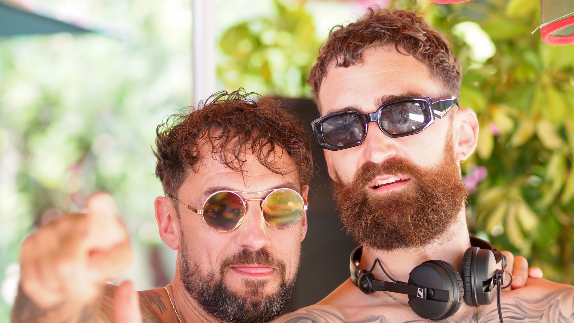 Two men wearing sunglasses pose closely together outdoors in bright sunlight.