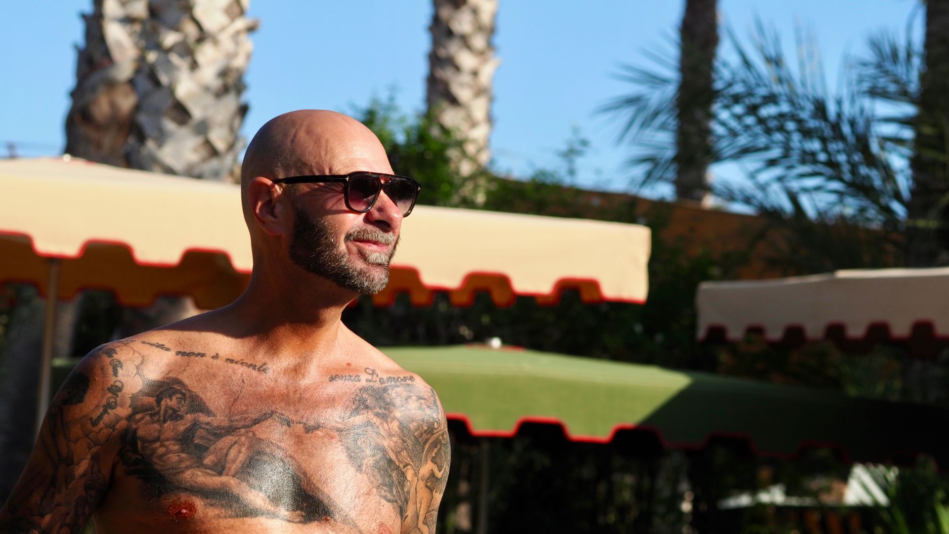 Shirtless man wearing sunglasses, standing outdoors in the sun with palm trees and awnings behind him.
