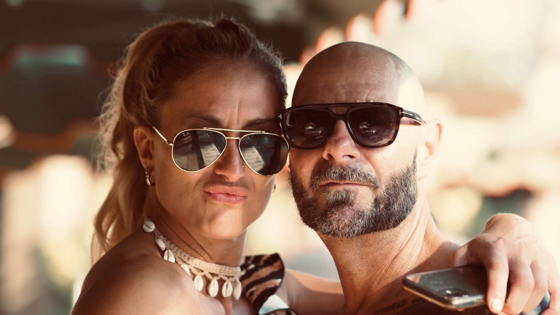 Two adults wearing sunglasses pose closely together outdoors in warm sunlight.