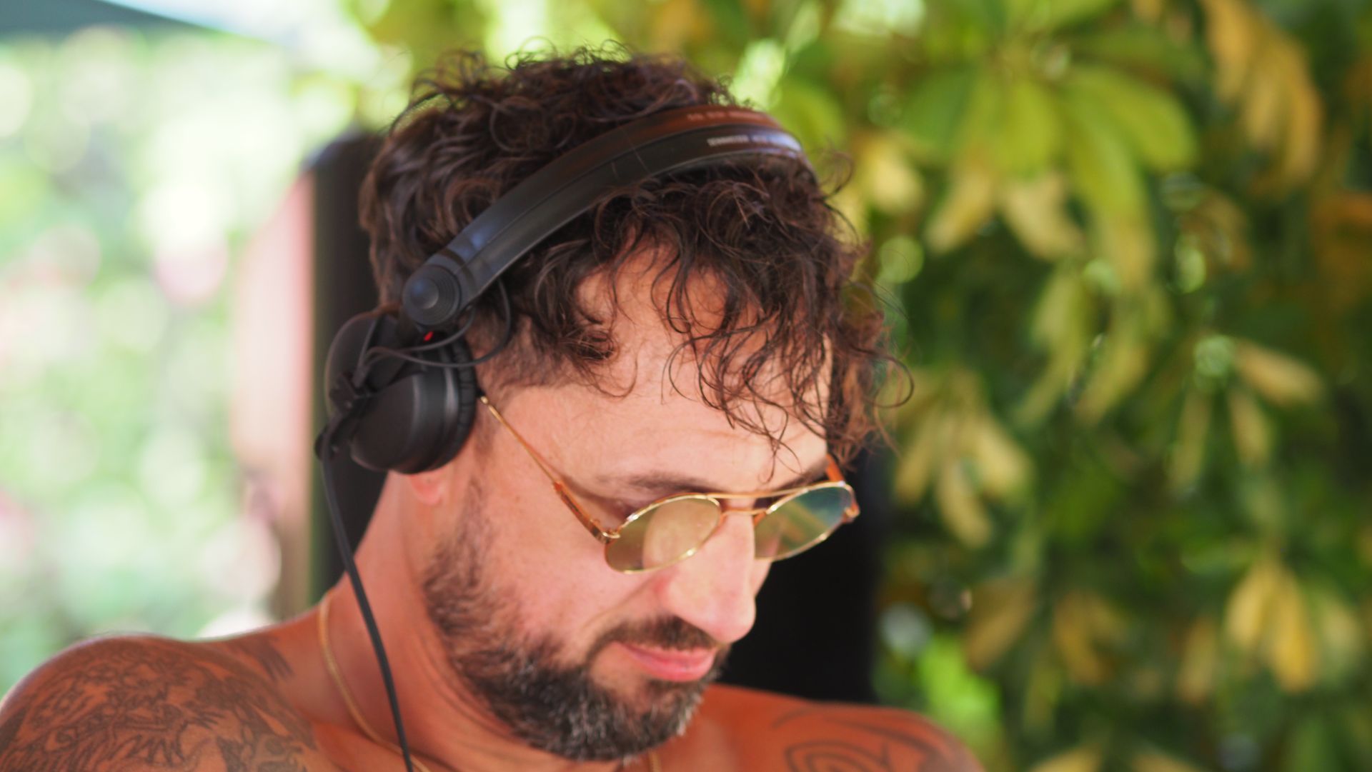 Man wearing headphones and yellow-tinted glasses looking down against a leafy green background