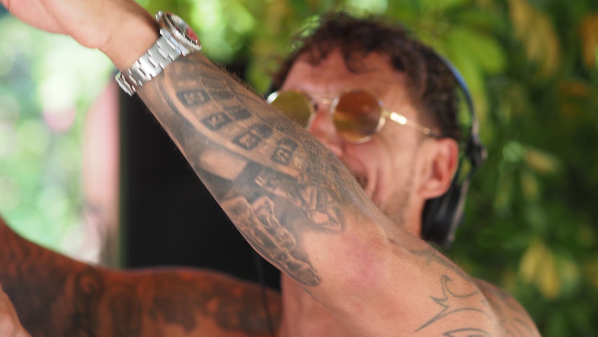 Tattooed person wearing sunglasses, raising an arm outdoors with blurred greenery in the background