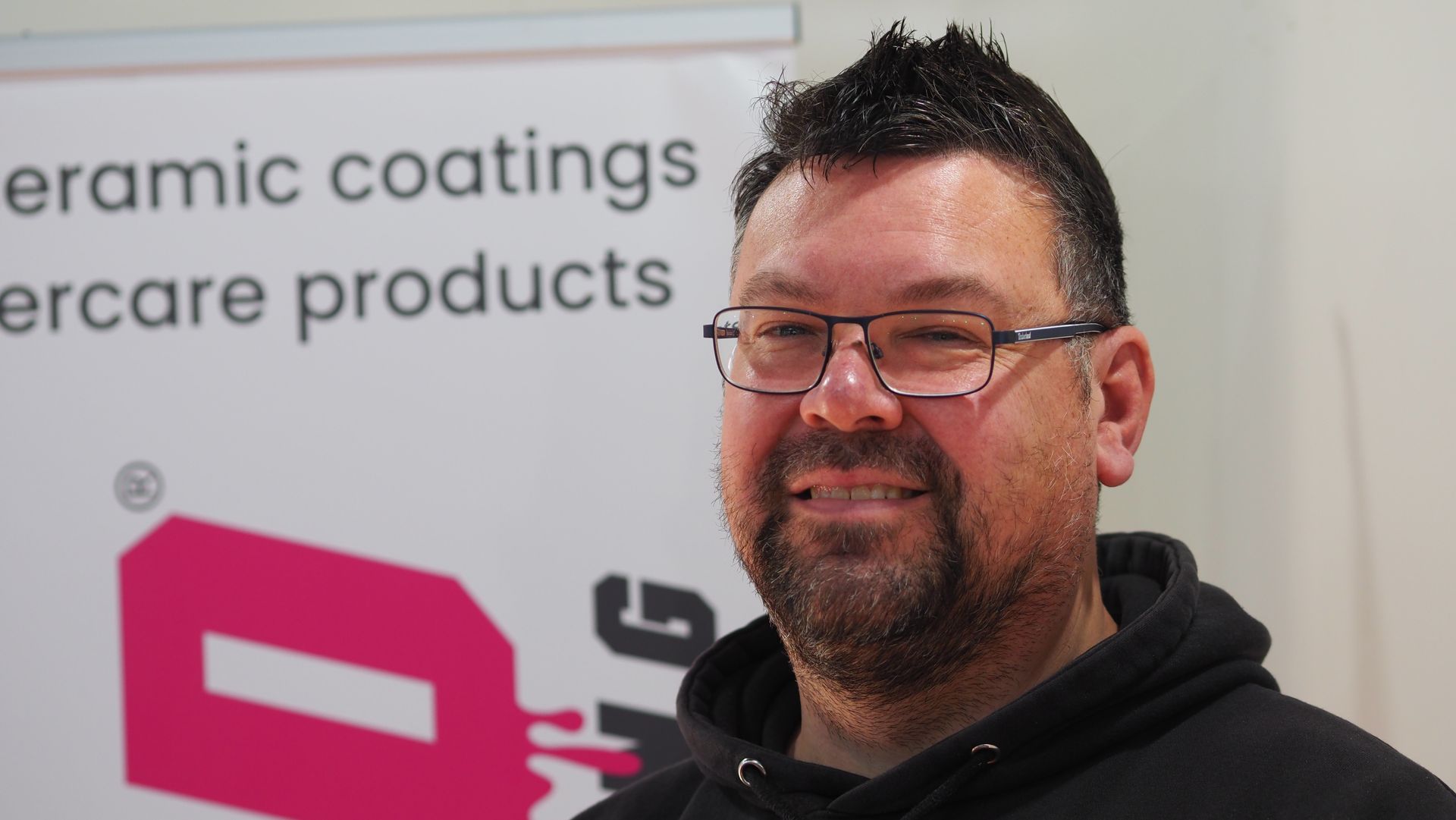 Smiling man with glasses in a black hoodie standing before a ceramic coatings trade show sign