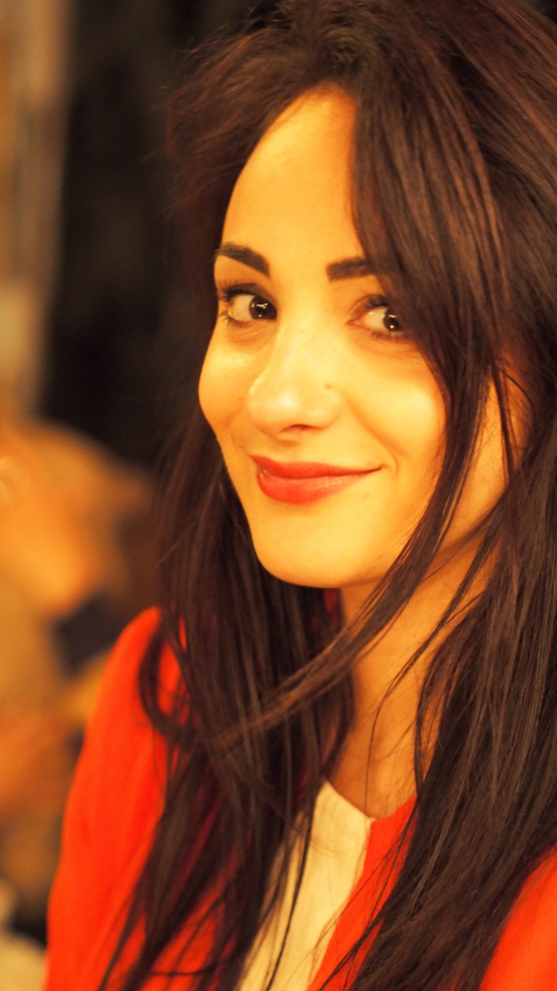 Smiling woman with long dark hair in a red top, warmly lit against a blurred background