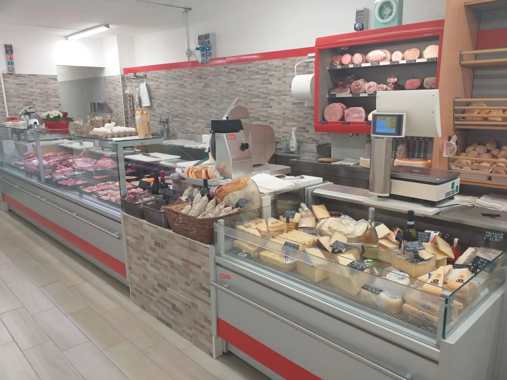 Un banco gastronomia con una vetrina piena di formaggi assortiti, accanto a reperti di salumi e una bilancia su uno scaffale sul retro.