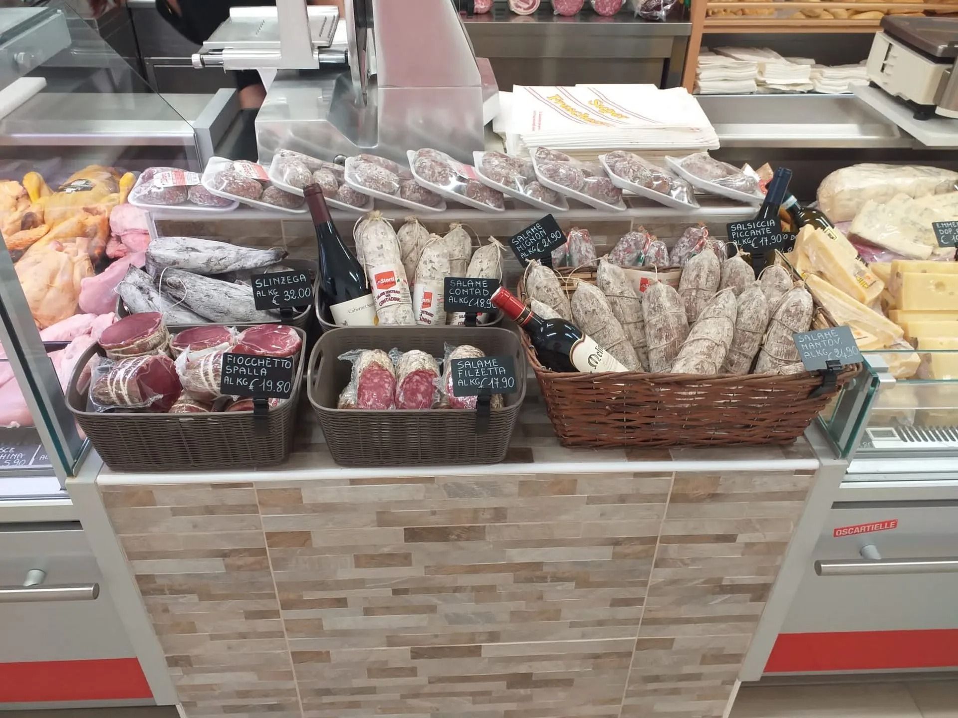 In una vetrina gastronomica sono esposti cestini e vassoi pieni di salsicce e salumi di vario genere, accanto a bottiglie di vino.