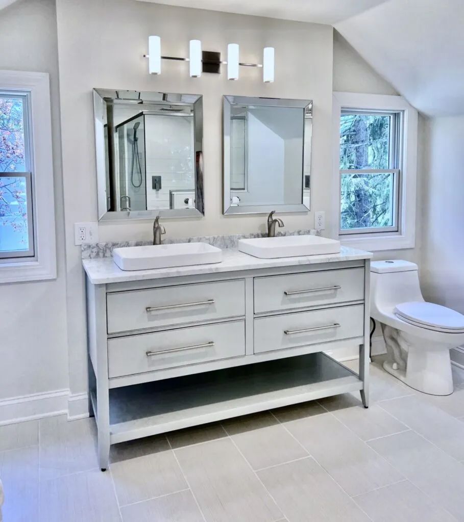 Bright white bathroom with double vanity, mirrors, windows, and a toilet beside the sink area.