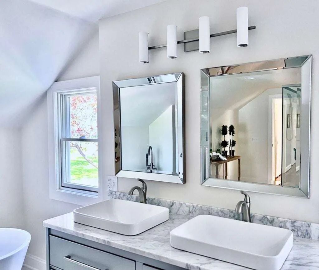 Bright white bathroom vanity with double sinks, mirrors, wall lights, and a small window.
