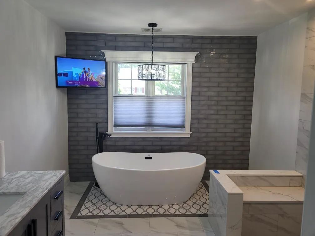 Modern bathroom with freestanding tub, gray tile accent wall, wall-mounted TV, and window with blinds