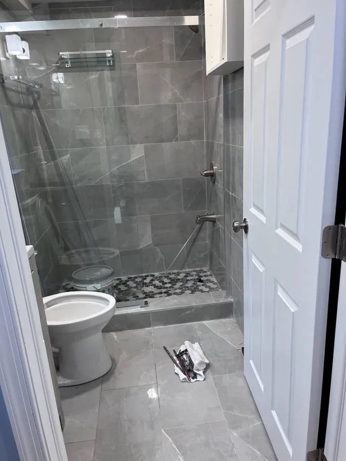 Small bathroom with gray tile, glass shower, toilet, and an open door.
