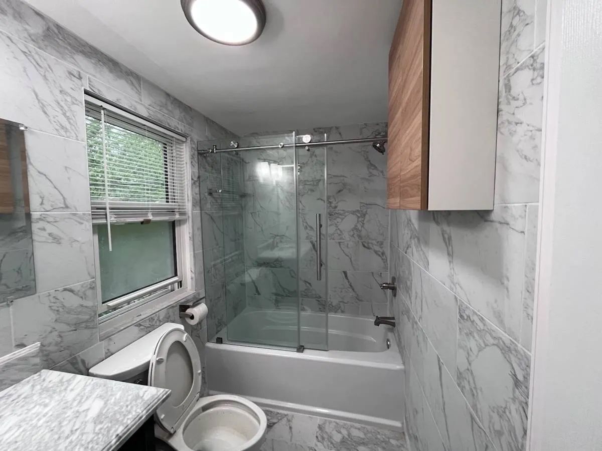 Small bathroom with marble walls, bathtub-shower combo, toilet, window, and wooden cabinet.