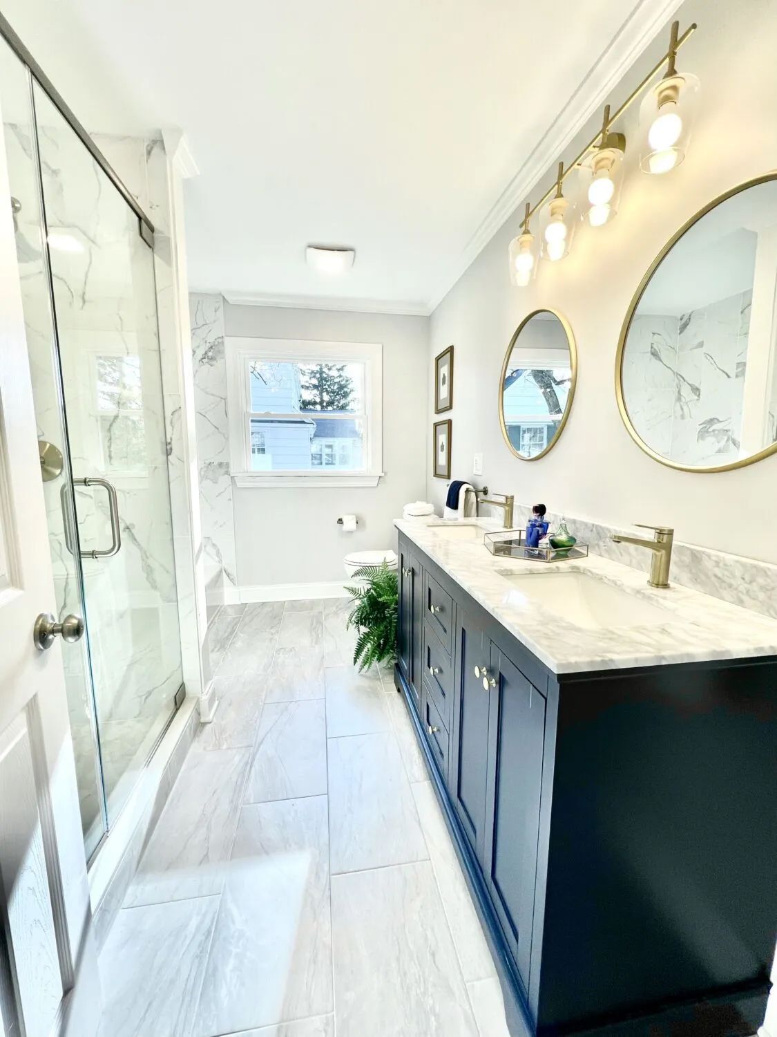 Bright modern bathroom with glass shower, double vanity, round mirrors, and large window.