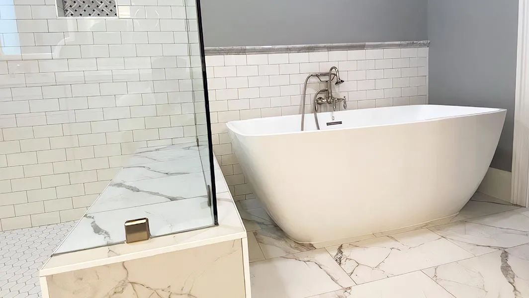 Modern bathroom with a white freestanding tub, glass shower panel, and marble tile floor and wall.