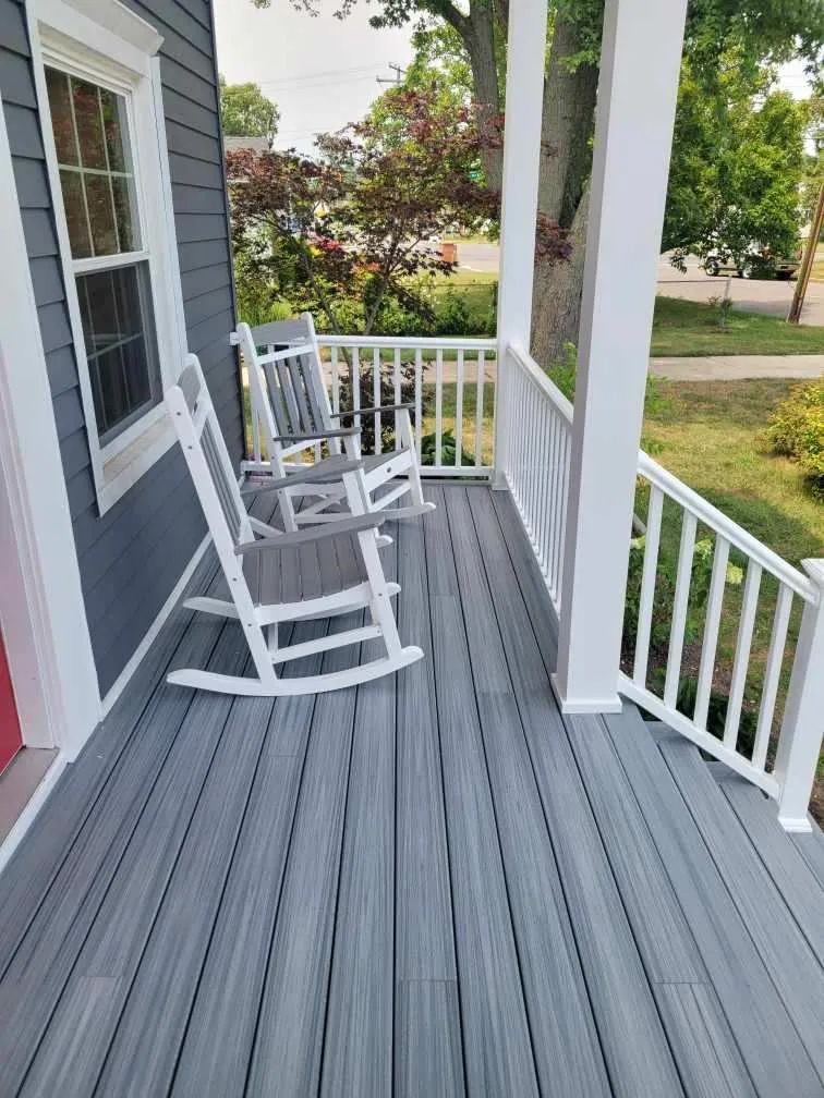 Front porch with gray rocking chair, white railing, and red house siding in a sunny yard