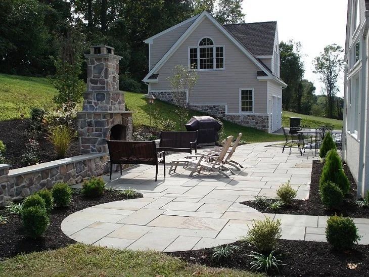 Stone patio with outdoor seating and fire pit beside a gray house and chimney in a landscaped backyard