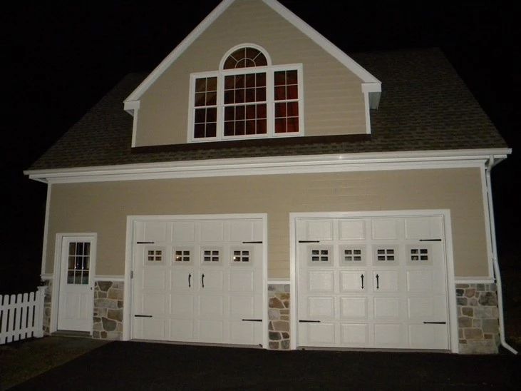 Two-story beige garage with white double doors and dormer window at night