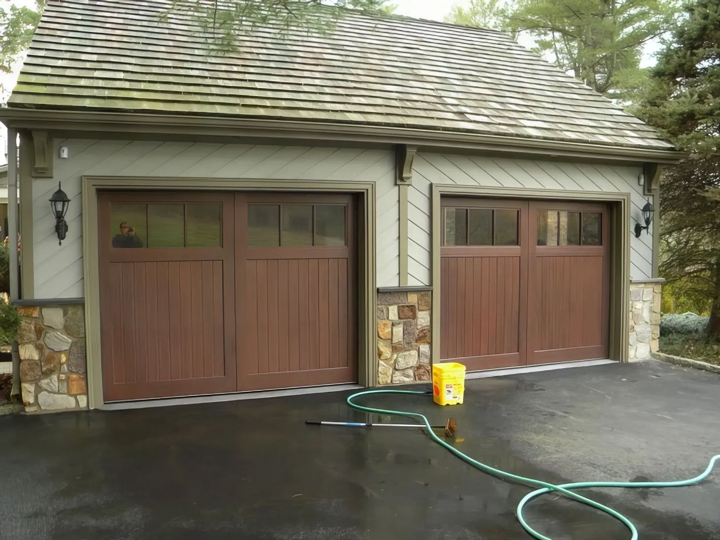 Two-car garage with brown doors, stone trim, and a green hose on a wet driveway.