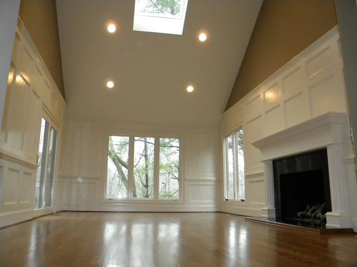 Empty beige living room with high ceiling, skylight, large windows, and a fireplace