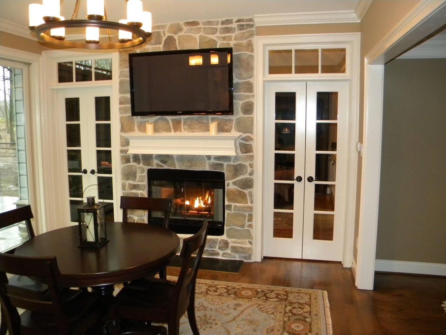 Dining area with round table, fireplace, wall-mounted TV, and French doors in a stone-accented room