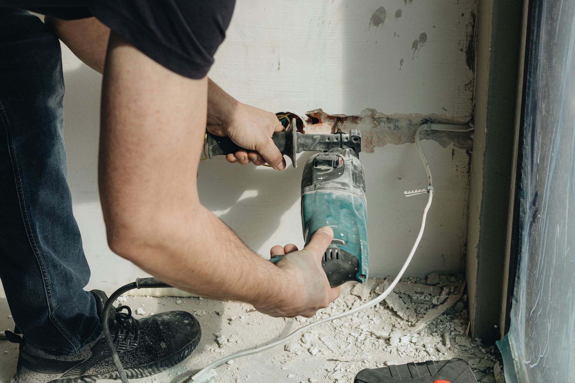 Person drilling into a wall beside an electrical box, with dust and debris on the floor.