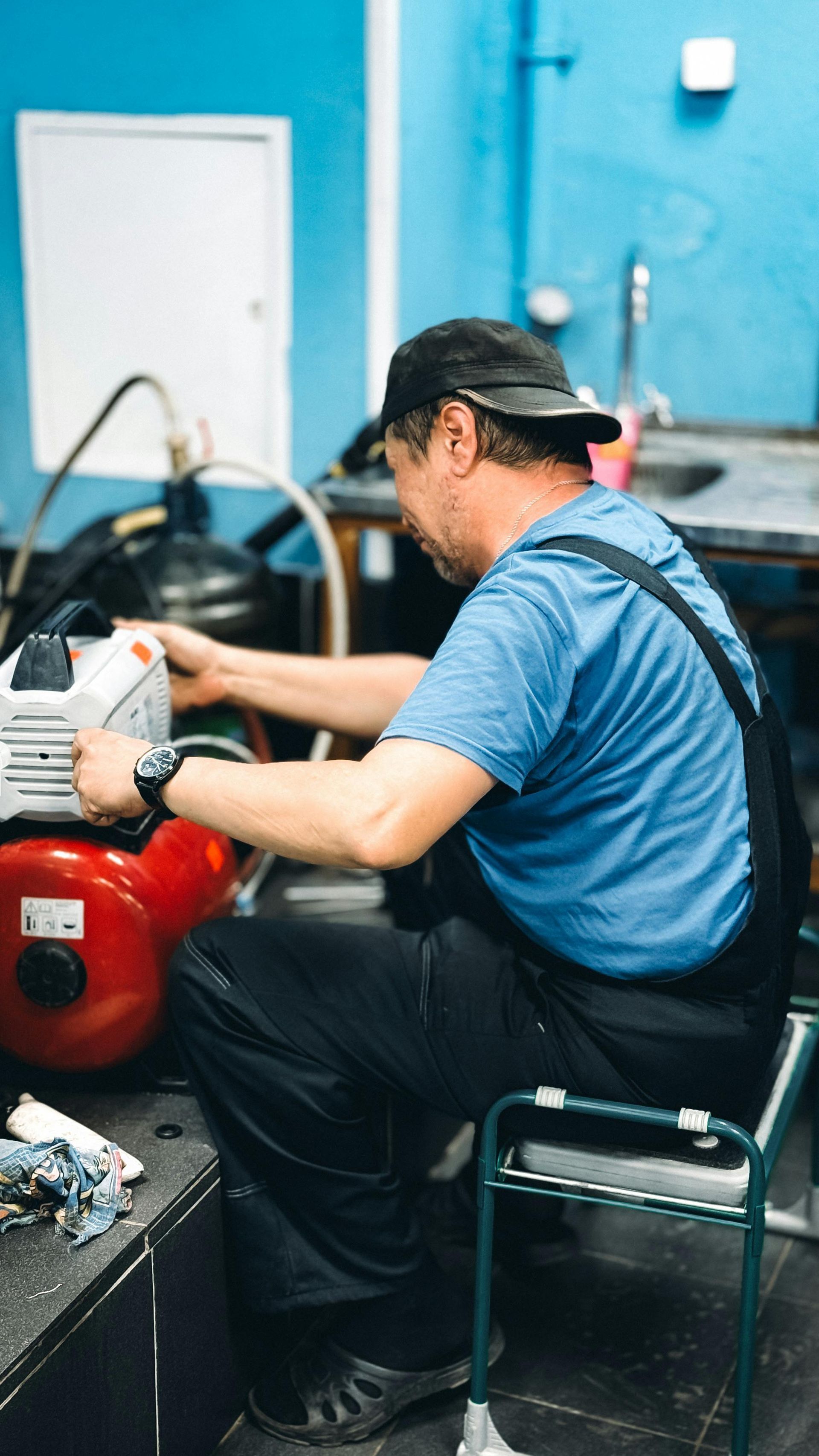 Worker in blue shirt operating a red machine outdoors beside a blue wall