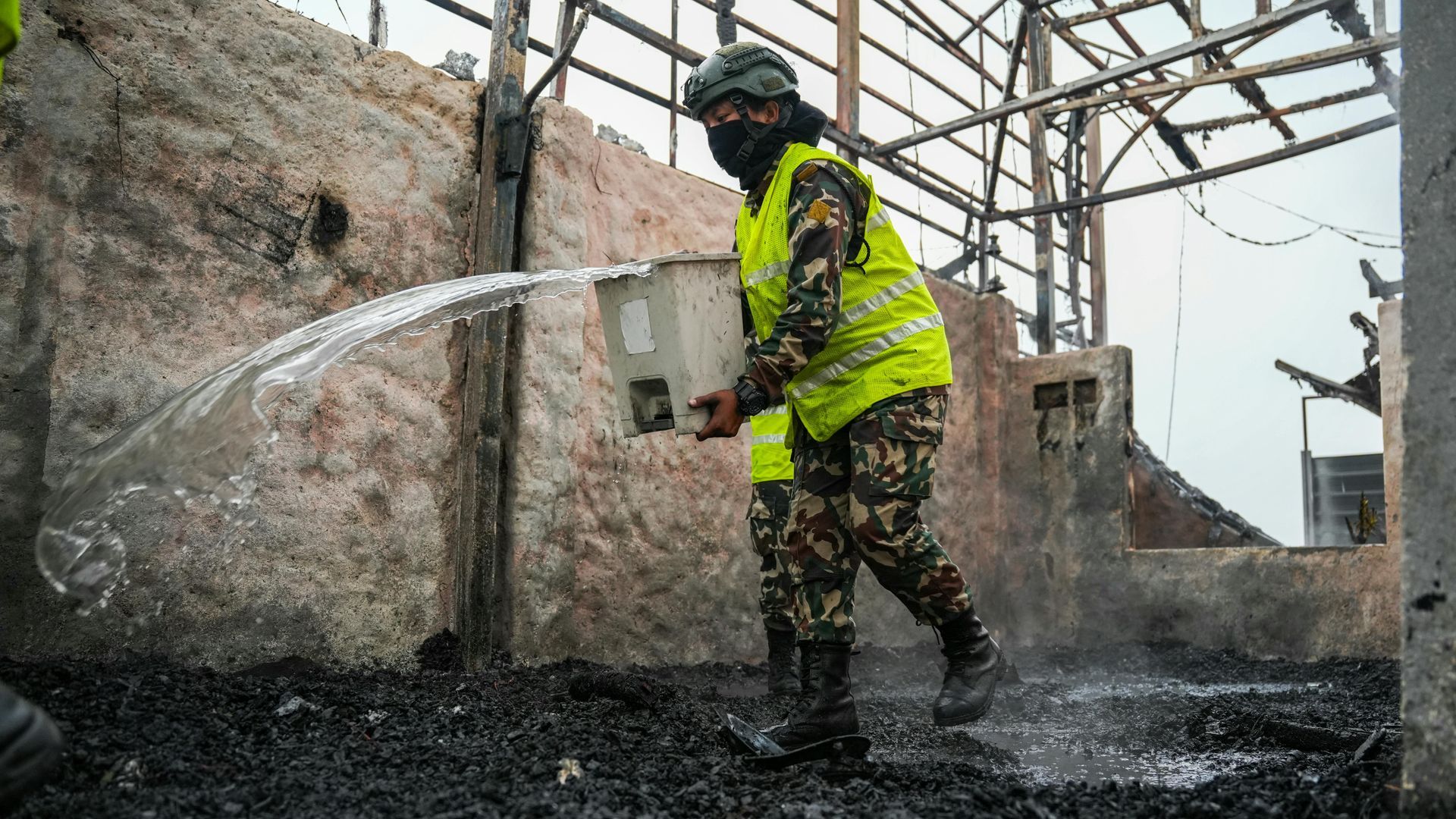 Firefighter in yellow gear spraying water at a smoky, damaged structure amid rubble.