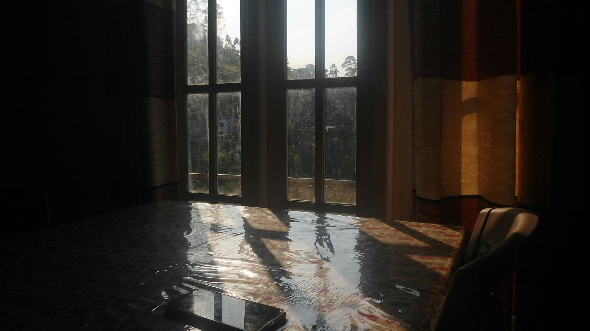 Sunlit dining table by tall windows with reflections and a chair in a dim room