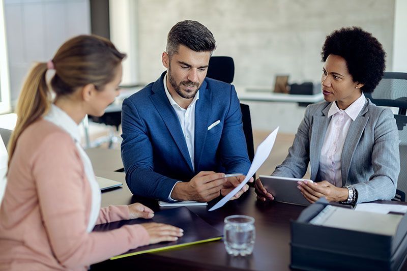Three professionals in a modern office, two sitting at a table with documents and a tablet, one facing them.