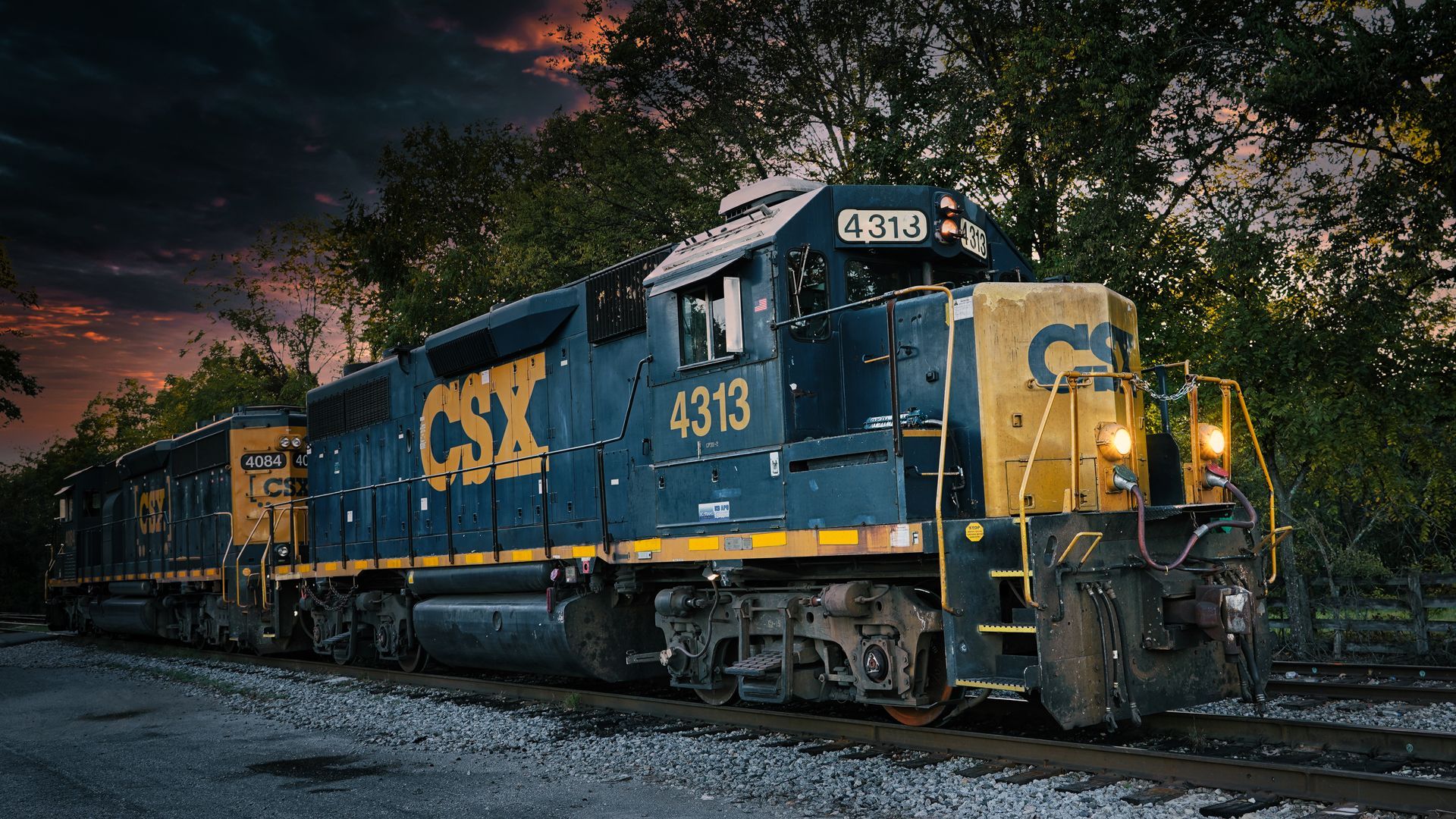 Blue CSX freight locomotive on tracks at dusk, with trees and dark sky in the background