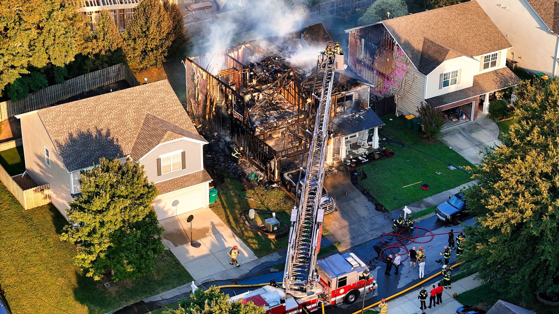 Fire-damaged house with firefighters battling flames from a ladder truck in a suburban neighborhood
