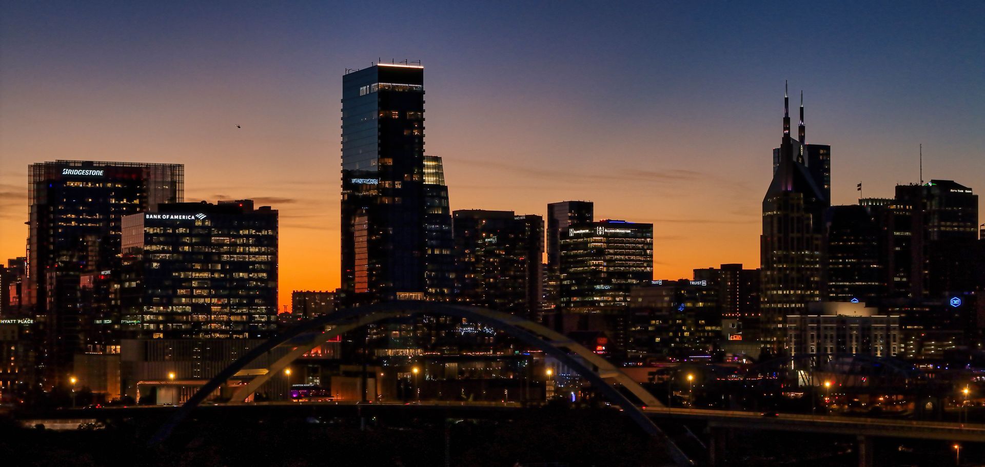 Nashville city skyline at dusk with lit skyscrapers and a bridge silhouetted against an orange-blue sky