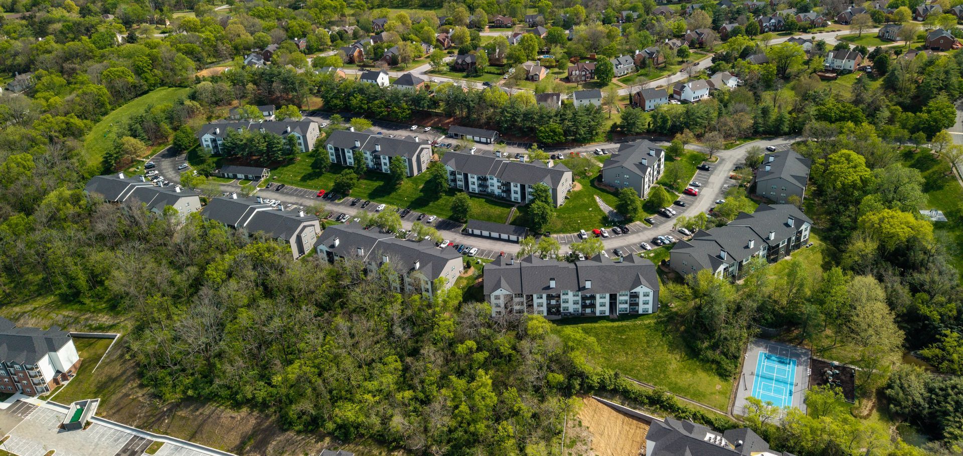 Aerial view of a tree-lined apartment complex with gray rooftops and a blue swimming pool
