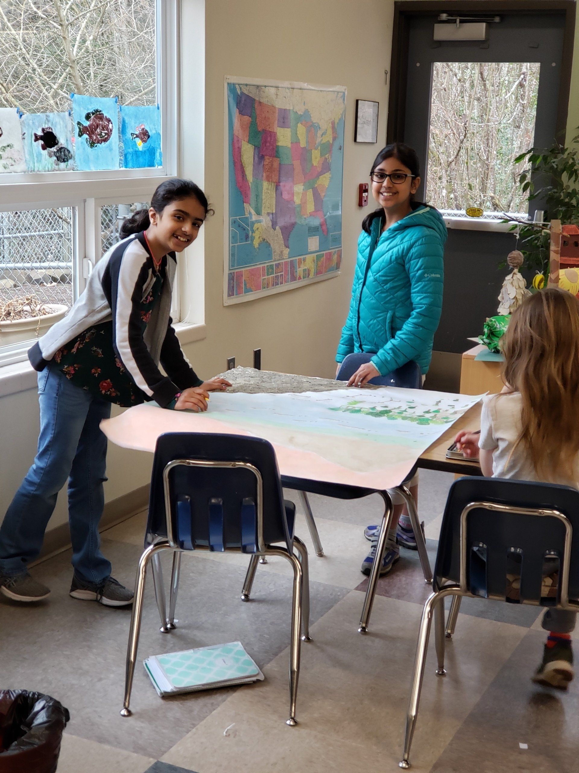 A group of adolescents are standing around a table with a map of the united states on the wall.
