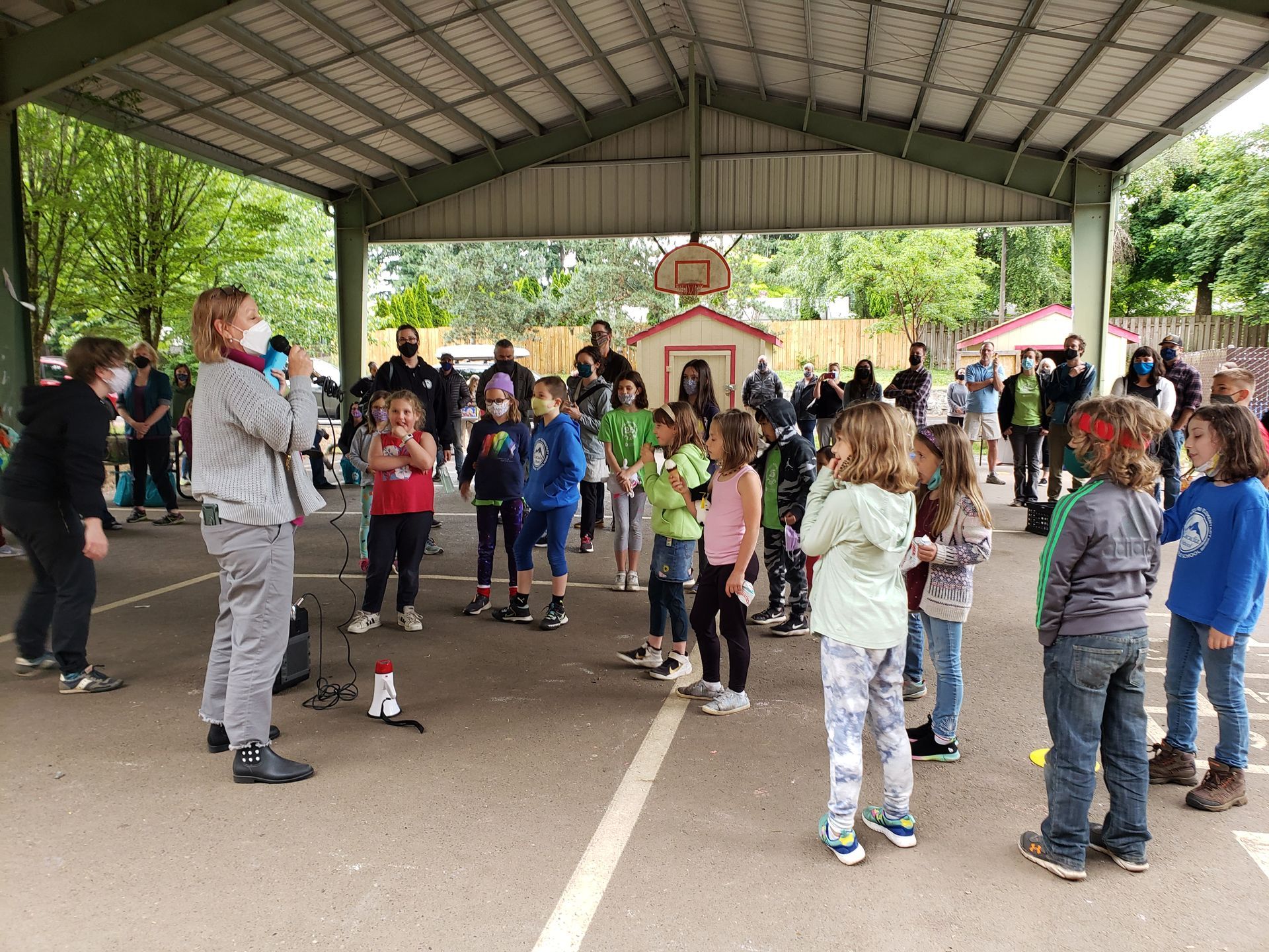A group of children are standing under a canopy in a playground listening to a coach