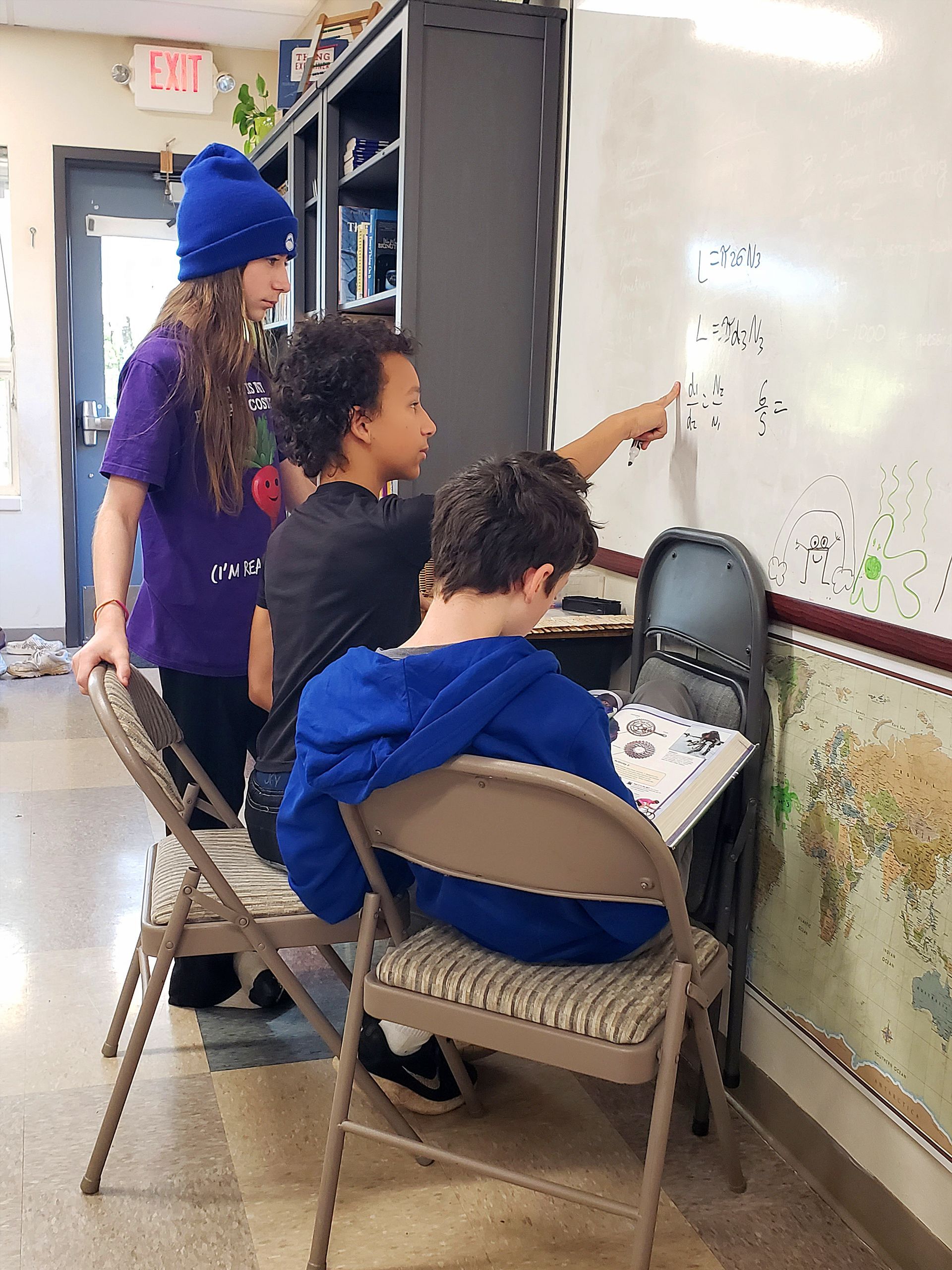 A group of adolescents are sitting in chairs in front of a white board doing mathwork toghether