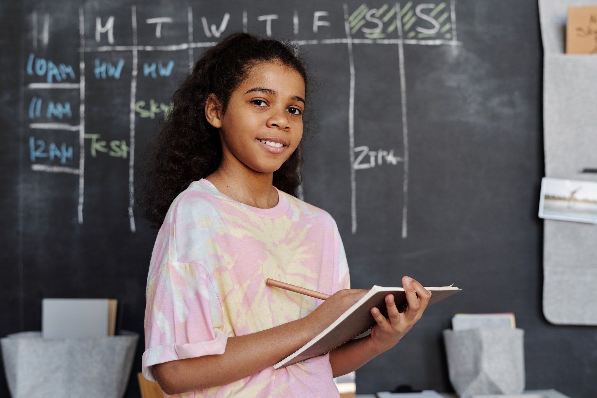 A young girl is standing in front of a blackboard holding a notebook and a pen.