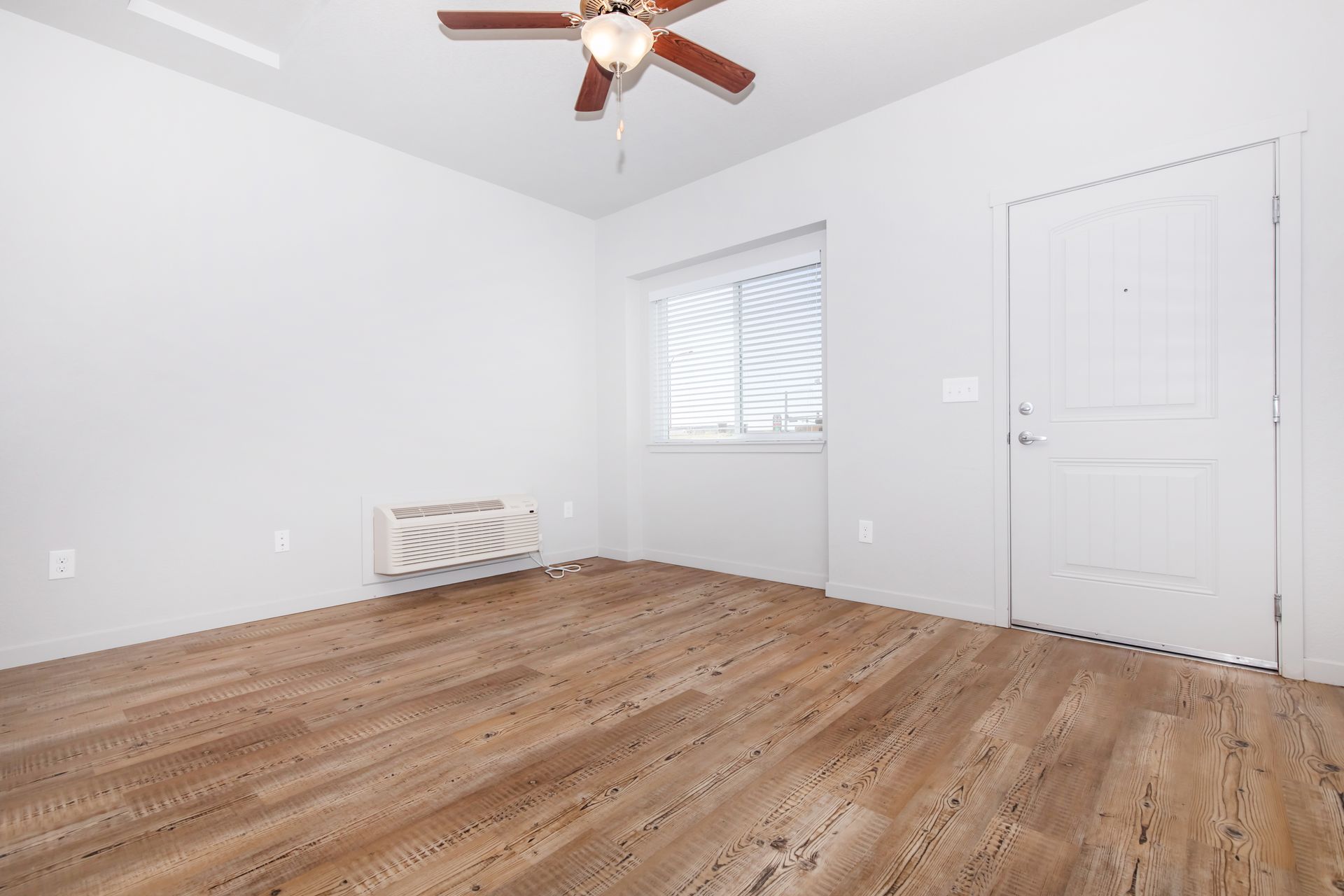 Empty white bedroom with wood floors, ceiling fan, window, and closed door