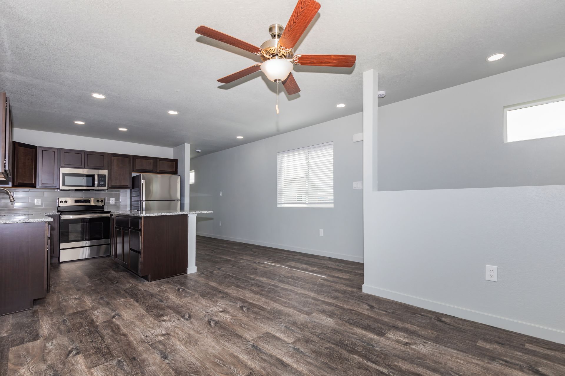 Empty open-plan kitchen and living room with gray walls, dark wood floors, and a ceiling fan