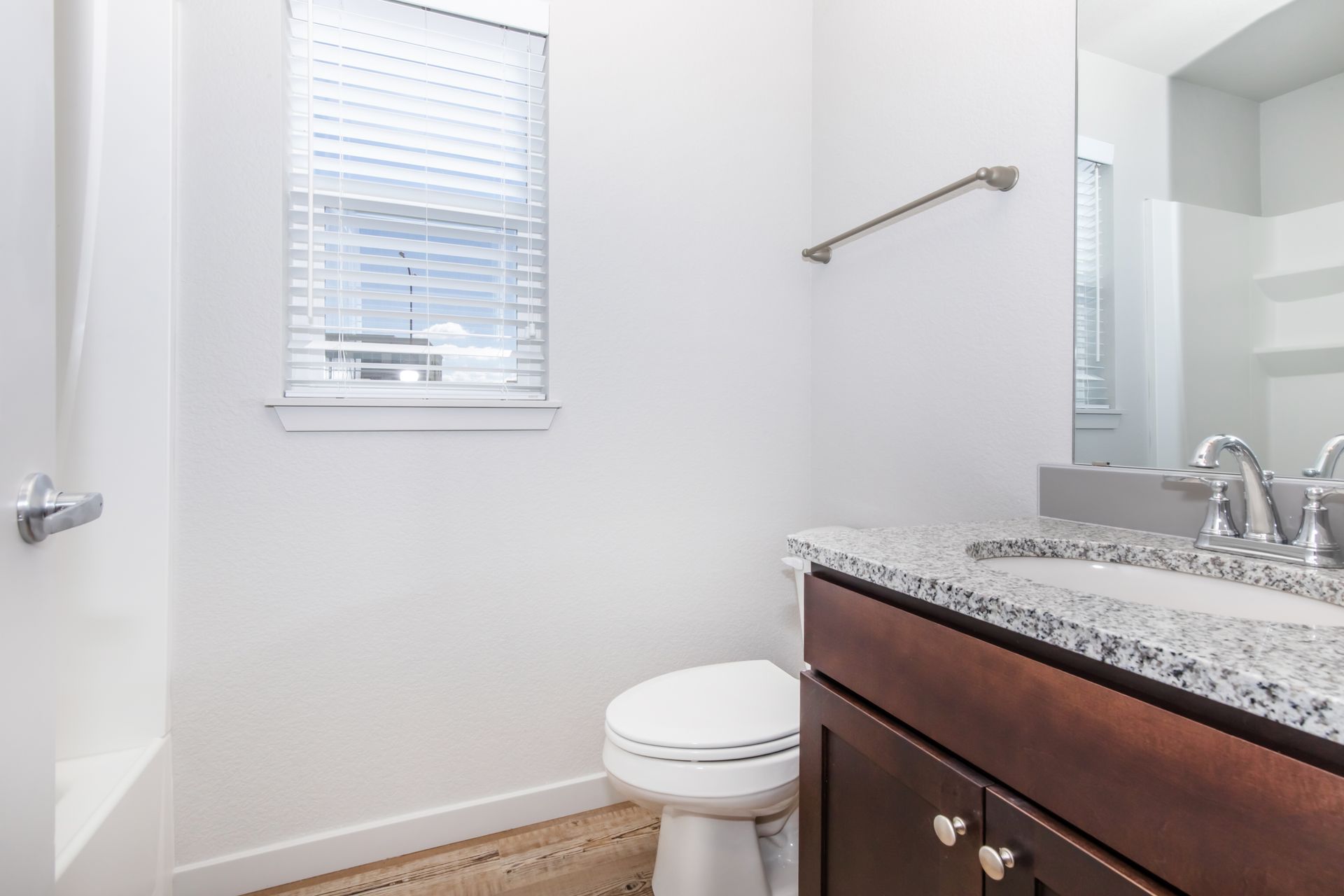 Bright bathroom with white walls, window blinds, toilet, and dark wood vanity with granite countertop.