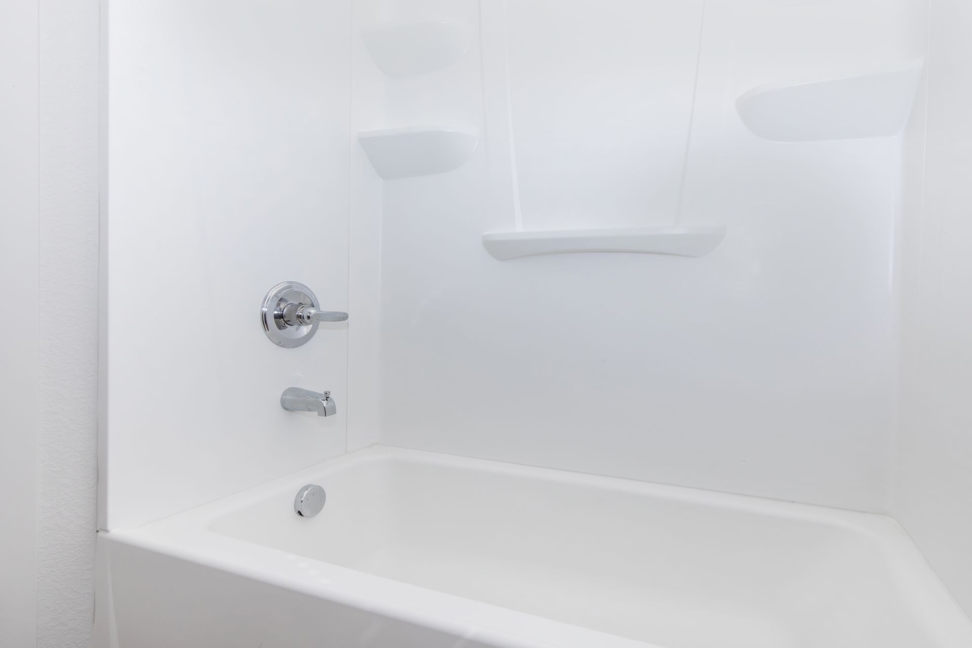 White bathtub and shower surround with chrome fixtures in a bright, empty bathroom.