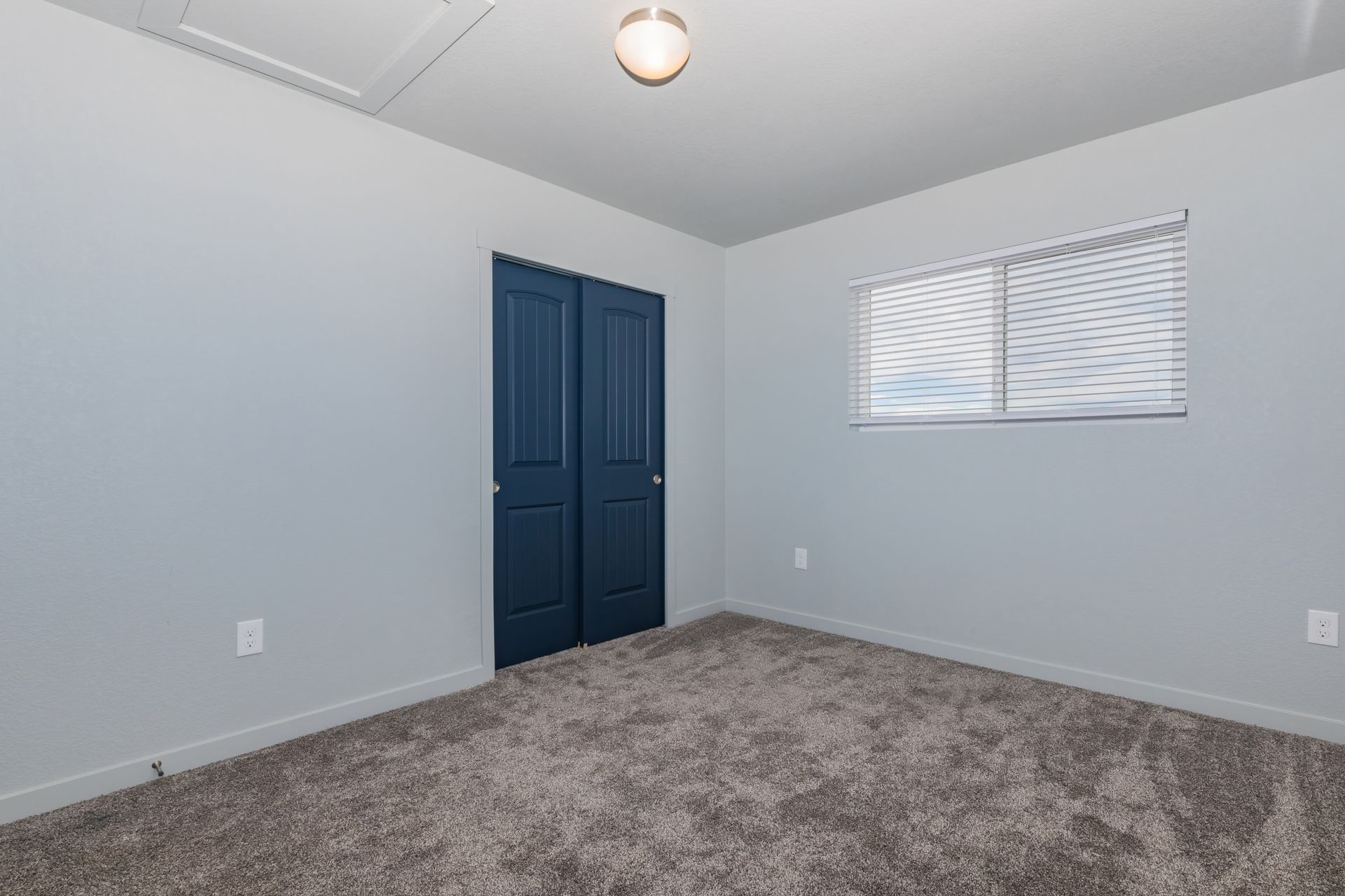 Empty bedroom with gray carpet, light gray walls, blue closet doors, and a window with blinds.