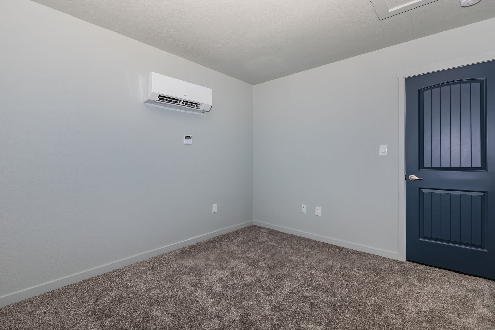 Empty gray carpeted room with white walls, wall-mounted unit, and a dark blue door