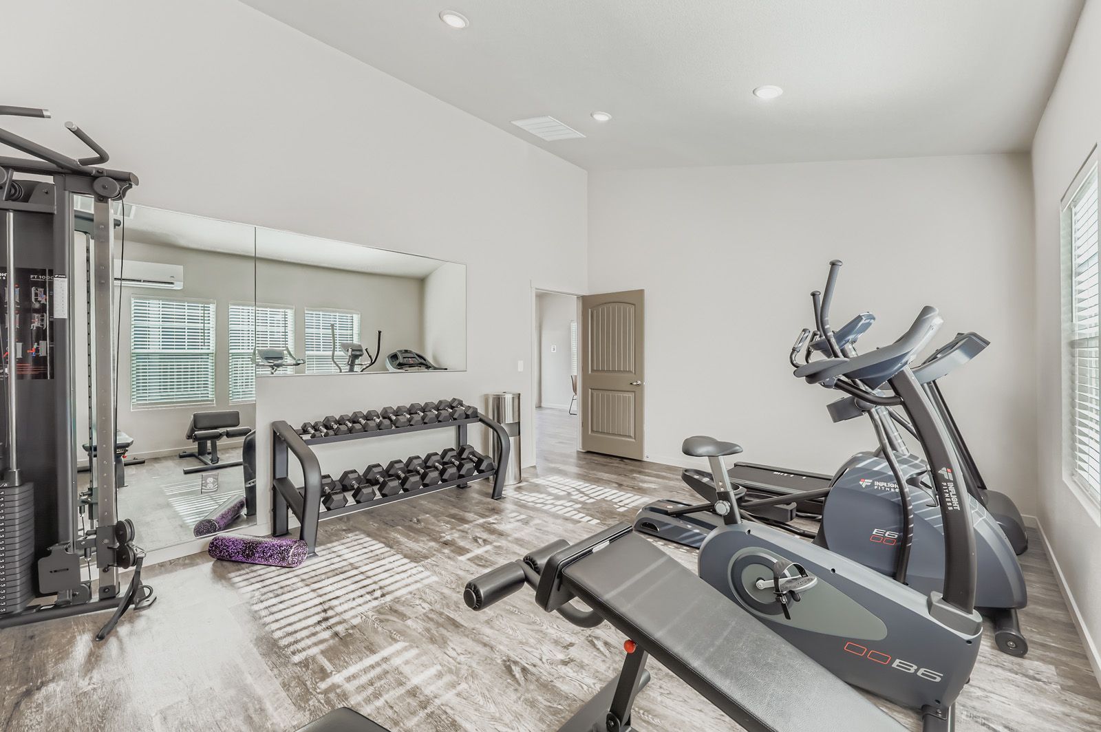 Modern home gym with treadmill, elliptical, weight machine, and dumbbell rack in a bright room