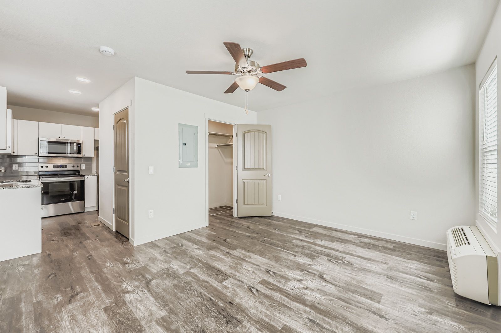 Empty modern apartment living area with wood-look floors, white walls, ceiling fan, and open kitchen.