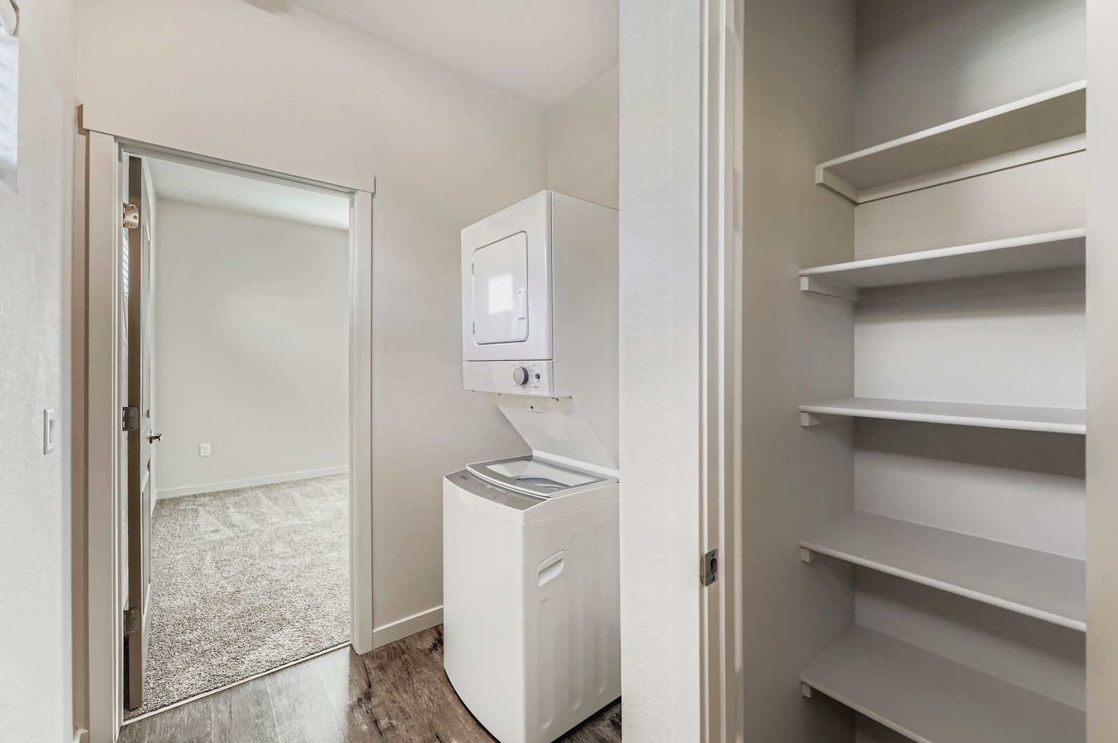 Laundry closet with stacked washer-dryer, white shelves, and an open doorway to a carpeted room.