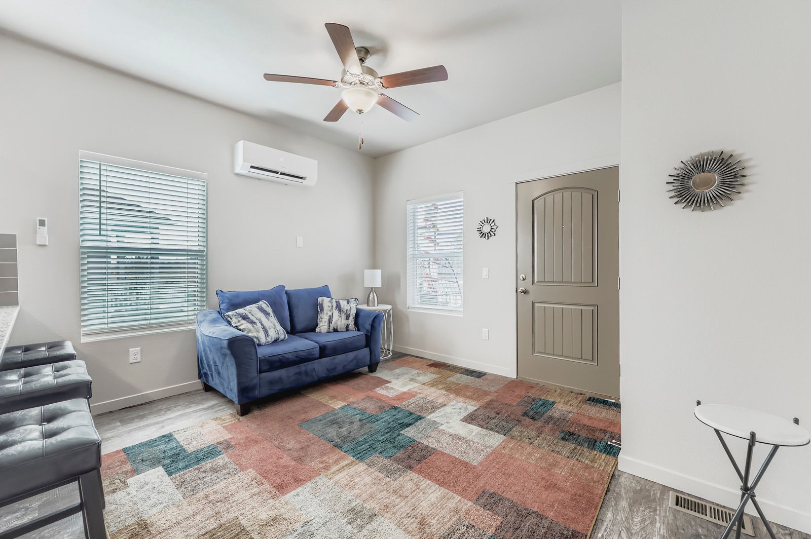 Bright living room with blue sofa, colorful rug, ceiling fan, wall AC, and windows with blinds.