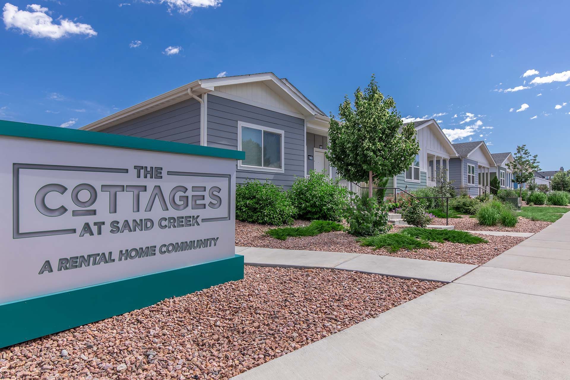 The Cottages at Sand Creek entrance sign beside a sidewalk and tan homes under a blue sky