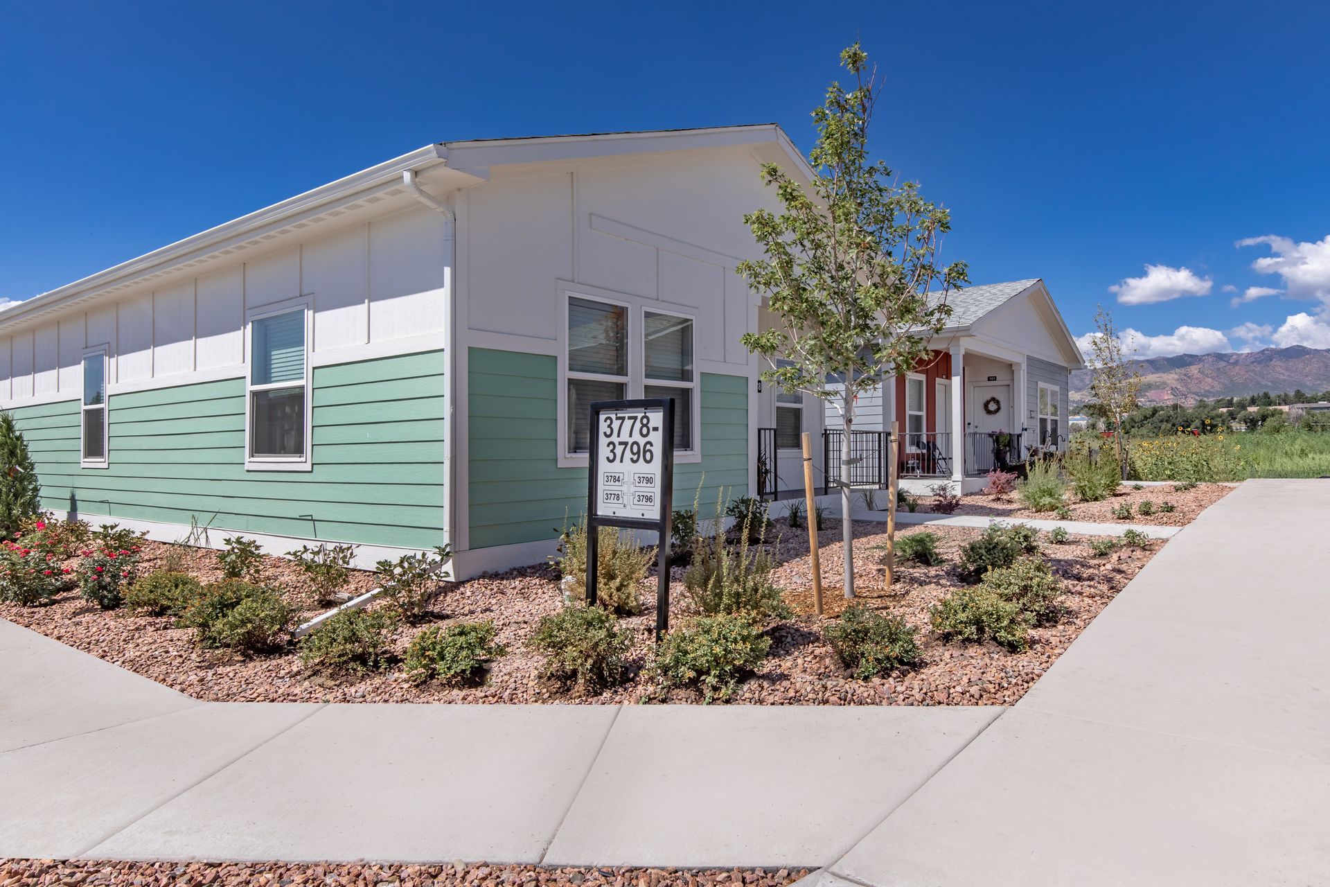 Modern single-story building with white and mint-green siding, a sidewalk, and a small tree under a blue sky