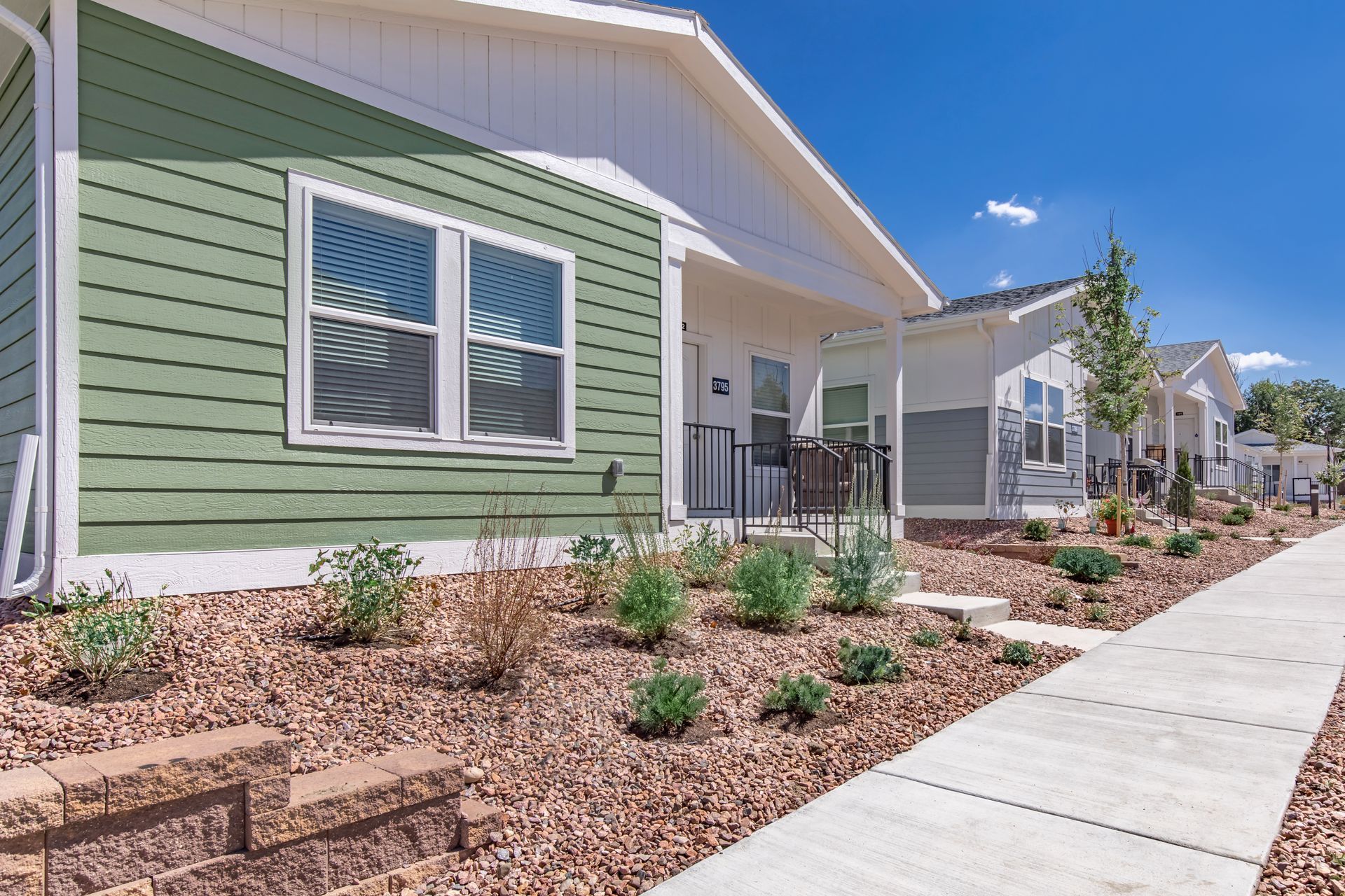 Modern single-story home with green siding, white trim, xeriscaped front yard, and a sidewalk under a blue sky